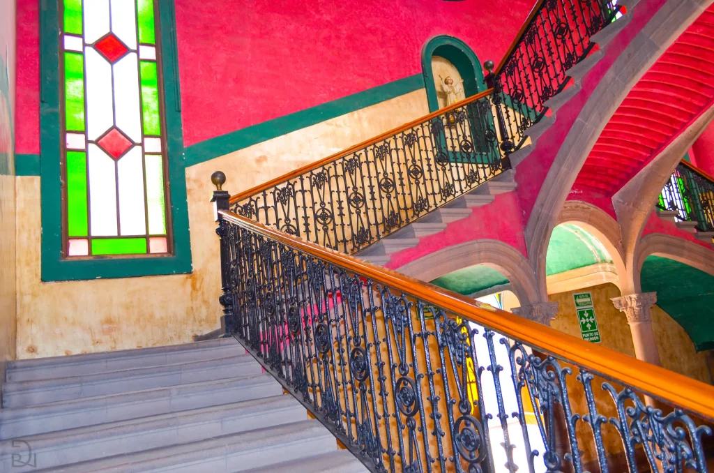 Escalera histórica con barandal de hierro forjado, muros rojos y vitral