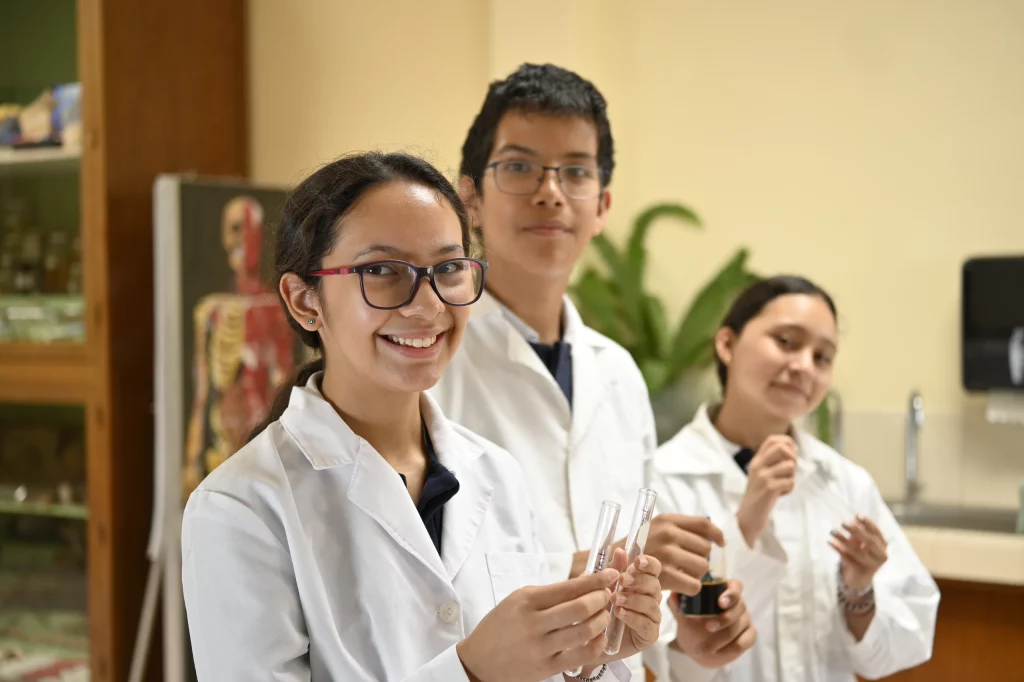 Tres alumnos con bata en el laboratorio, sonriendo a cámara