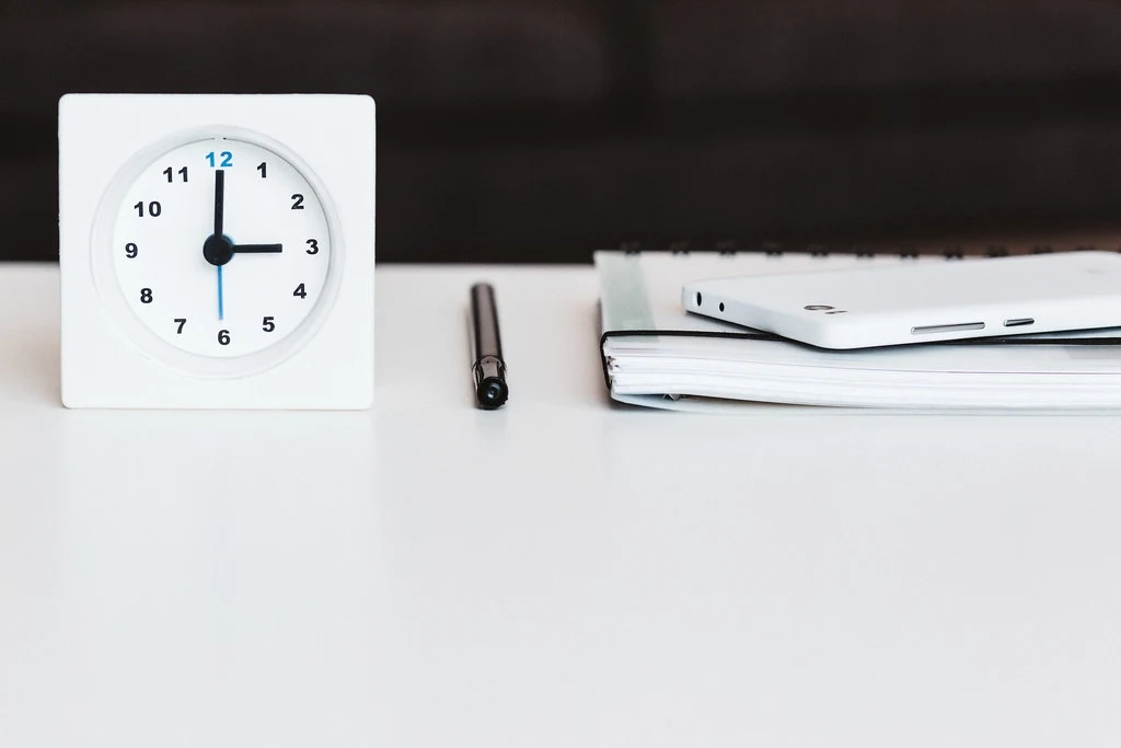Reloj de mesa, pluma y libreta sobre un escritorio blanco