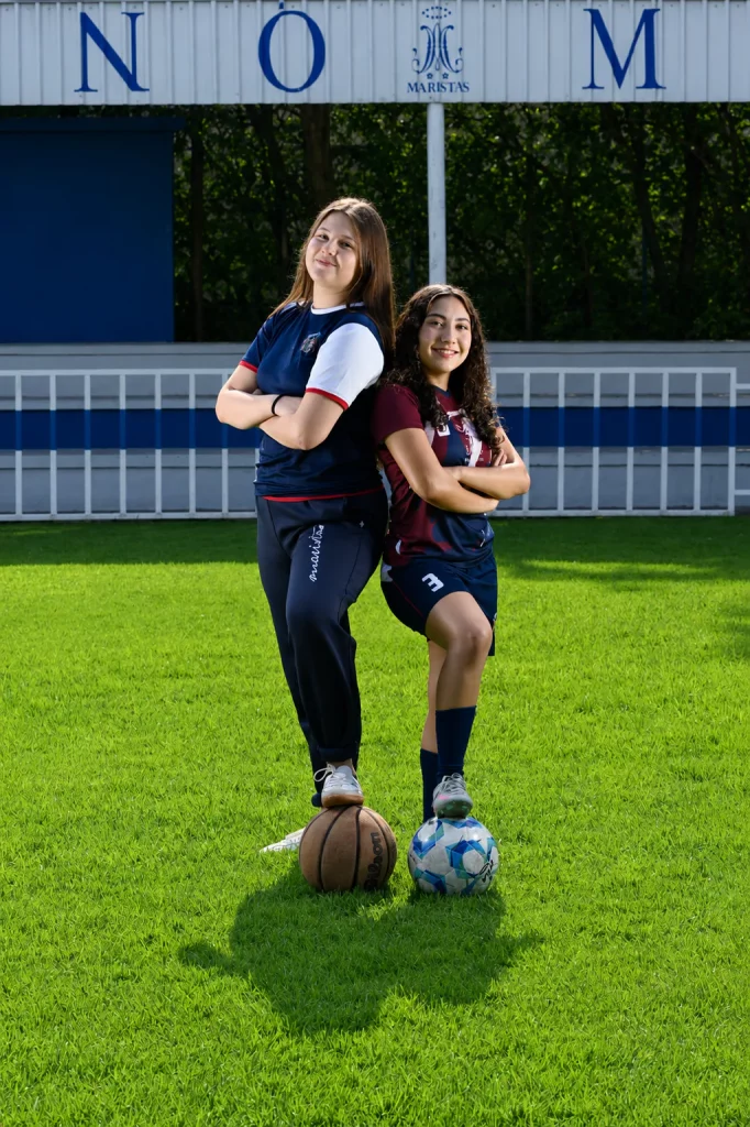 Dos alumnas con uniforme deportivo posando en el campo con balones